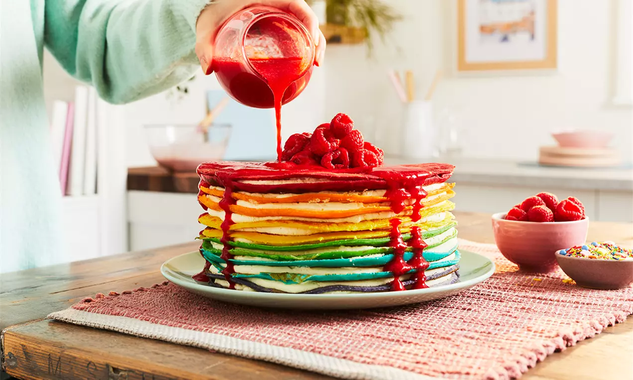 Tort stratificat din clătite colorate în nuanțe curcubeu, acoperit cu zmeură și sirop roșu, așezat pe o farfurie albă pe un șervet roz