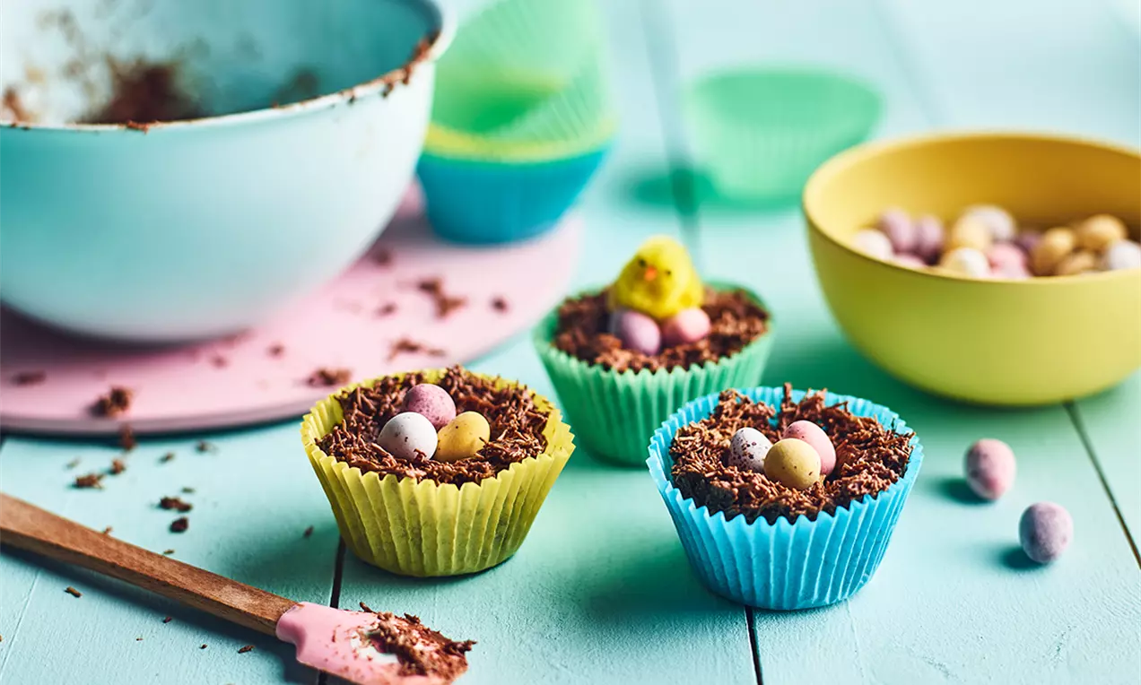 Cupcakes în forme colorate de hârtie, cuiburi din firimituri de ciocolată ce țin bomboane mici pastelate pe o masă albastră luminos decorată