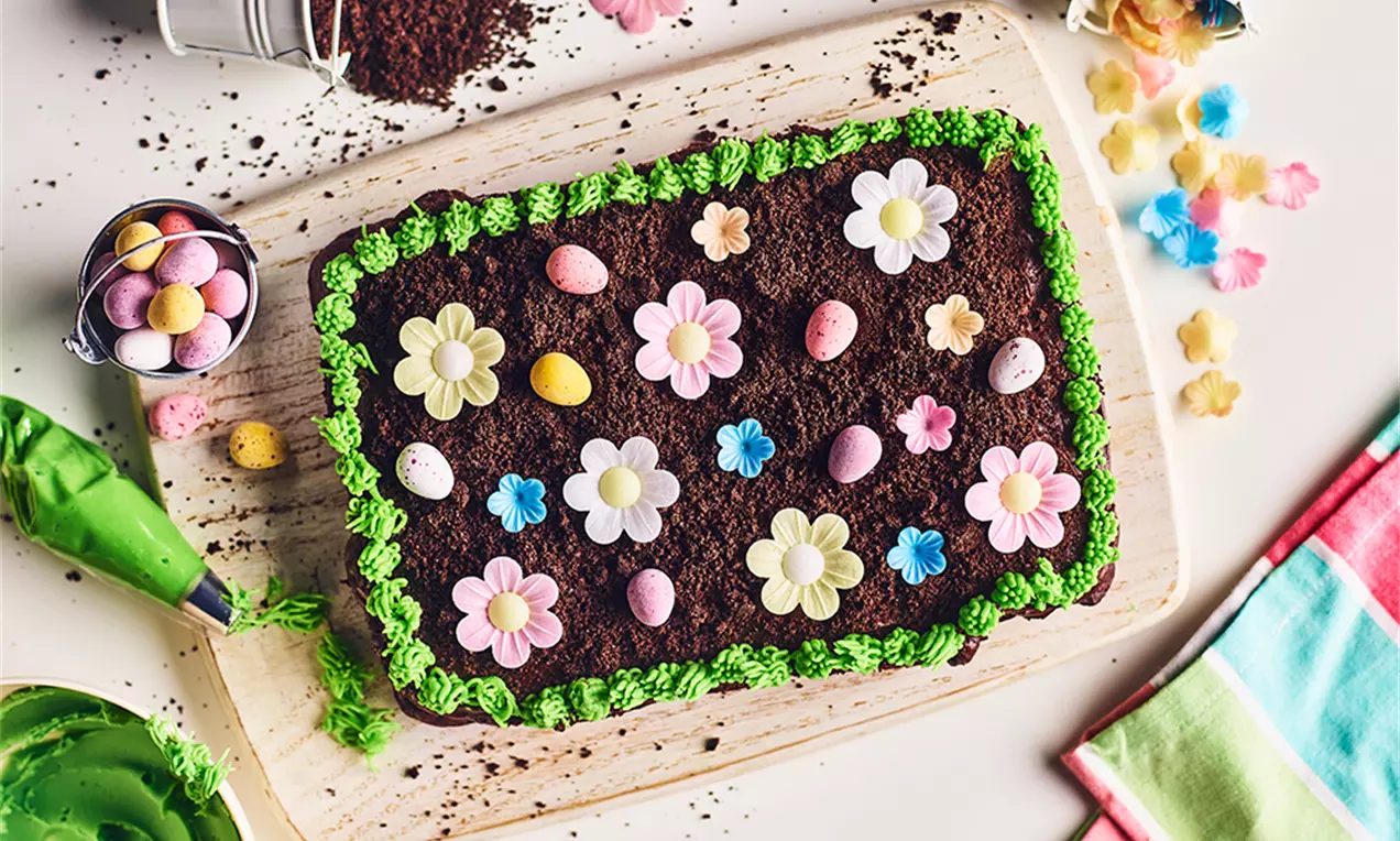 Prăjitură dreptunghiulară cu strat de cacao maroniu decorată cu flori multicolore și ouă pastelate pe bordură de cremă verde viu