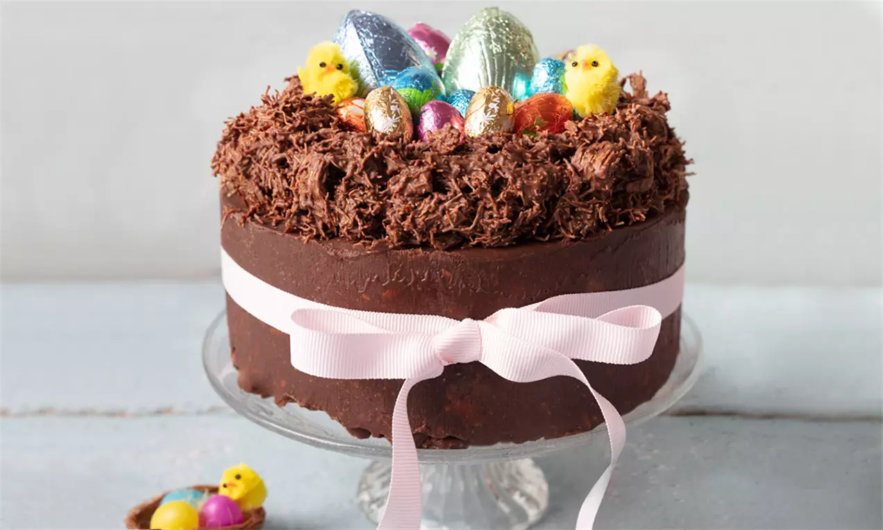 Tort rotund cu glazură lucioasă de ciocolată îmbrăcat într-o fundă roz, decorat cu un cuib din fulgi de ciocolată și ouă colorate