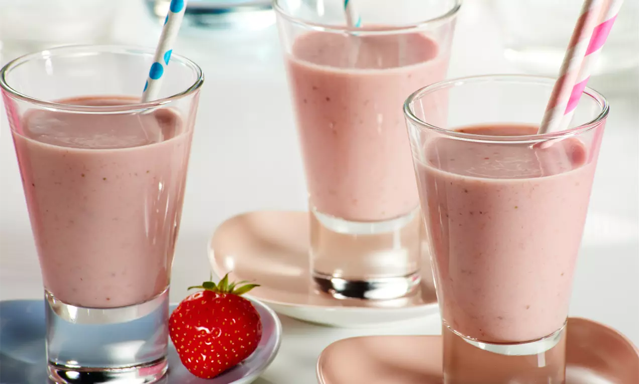 Milkshake roz-rozaliu din căpșuni, servit în pahare înalte cu pai colorat, însoțit de căpșuni întregi și farfurii pastelate asortate