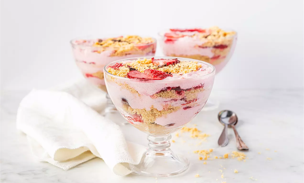 Parfait vegan cu straturi roz pal cremoase, căpșuni roșii vii și pesmet auriu, servit în cupă transparentă pe blat alb marmorat