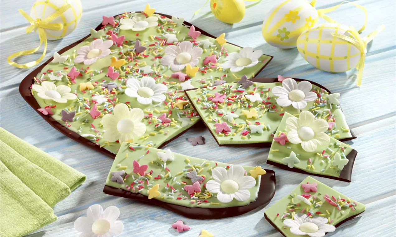 Placă subțire de ciocolată neagră acoperită cu glazură verde pastel, decorată cu flori albe, fluturi colorati și stropi multicolori