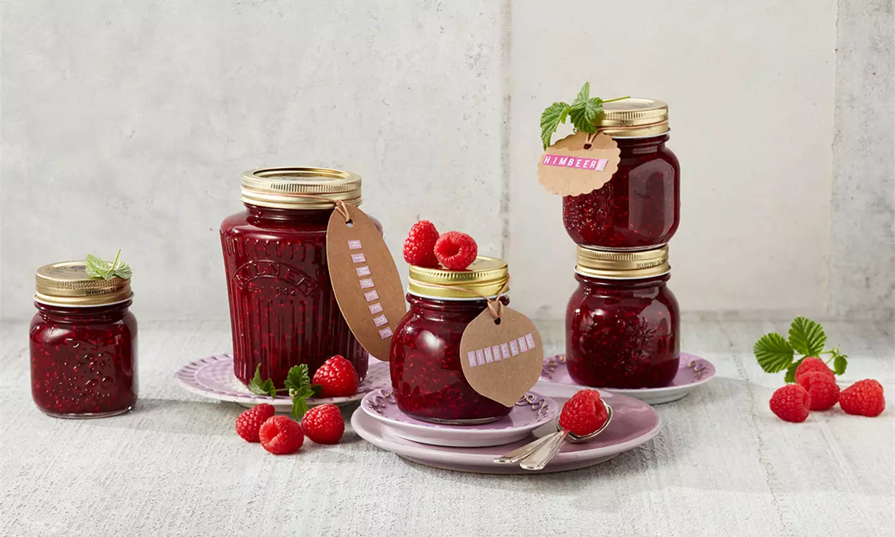 Borcane transparente cu dulceață densă de zmeură roșu-rubiniu, decorate cu etichete și frunze verzi, așezate pe farfurii pastelate cu zmeură proaspătă