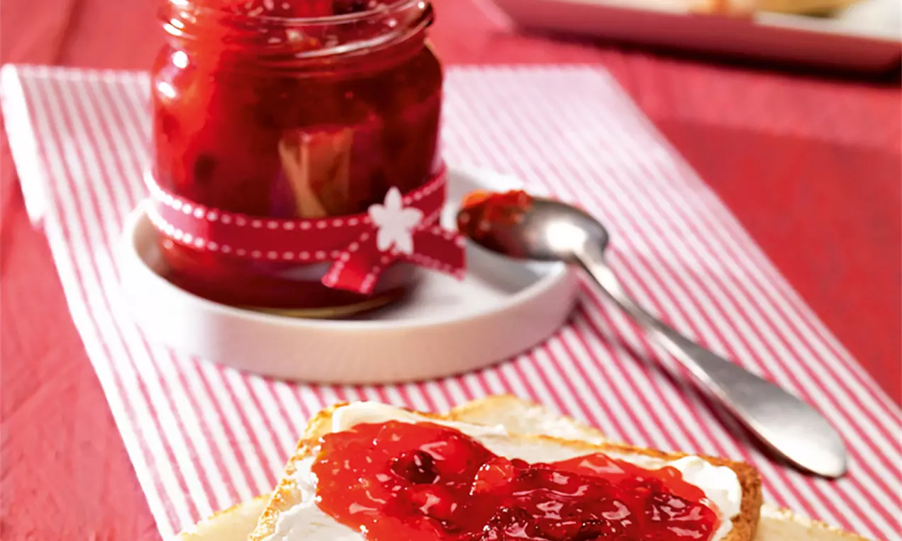 Dulceață roșu-rubinie cu textură densă, întinsă peste cremă albă pe pâine aurie, lângă un borcan decorat cu panglică și linguriță argintie