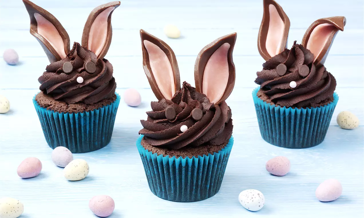 Cupcakes cu cremă de ciocolată maronie, decorate cu urechi din ciocolată și bomboane mici albe, așezate pe hârtii albastre și presărate cu ouă colorate