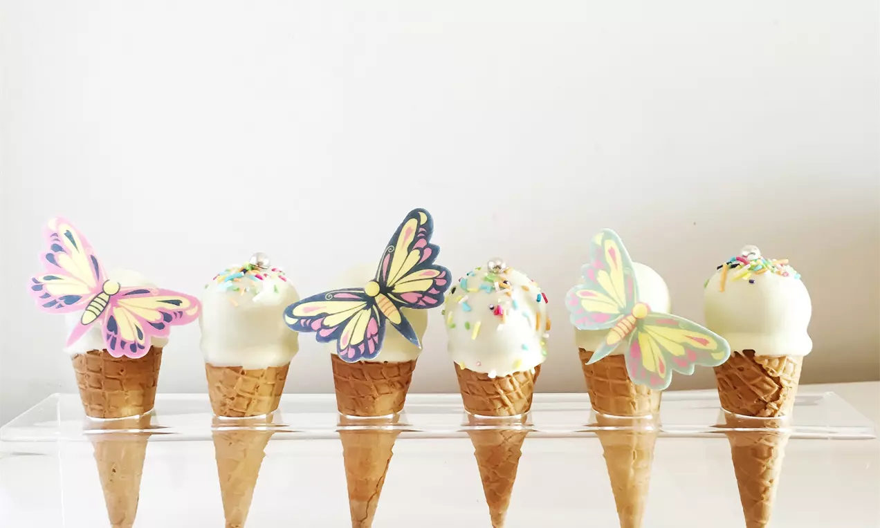 Înghețată albă-glazurată pe cornete aurii, decorată cu fluturi pastel și stropi colorati, expusă pe un suport transparent