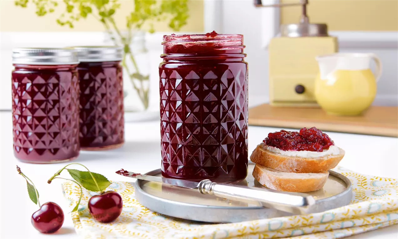 Dulceață de cireșe amare roșu-închis, într-un borcan cu model geometric, alături de felii de pâine unse și cireșe proaspete pe o farfurie argintie