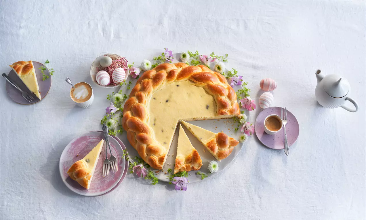 Pască rotundă cu aluat împletit auriu, umplută cu cremă de brânză vanilată și stafide, servită pe un platou decorat cu flori mov și albe