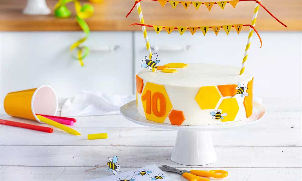 Tort rotund acoperit cu cremă albă, decorat cu faguri galbeni și portocalii, figurine de albine și banner colorat pe un suport alb