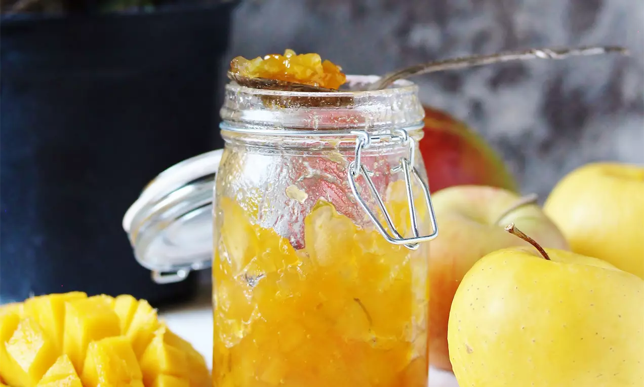 Dulceață aurie cu textură ușor densă în borcan transparent, alături de mere galbene, cuburi de mango și crenguțe de rozmarin pe un fundal rustic